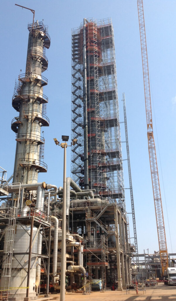 A large industrial plant with towering steel structures and scaffolding. A tall, cylindrical tower stands on the left, while a taller rectangular structure covered in industrial coatings is in the center. A crane extends into the blue sky on the right, highlighting the expertise of a commercial insulation company.