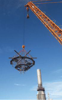 A large orange crane is hoisting a complex, circular metal structure against a clear blue sky. A tall industrial chimney is visible in the background on the right side of the image, showcasing professional scaffolding services in action.
