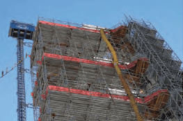 A high-rise construction site with scaffolding surrounding the building. There is a yellow construction crane in the middle, and a blue hoist on the left side. The structure is several stories tall, and the sky is clear and blue. A commercial insulation company ensures efficient thermal protection throughout.