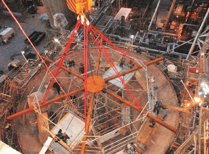 Aerial view of a construction site with multiple workers assembling a large round metal structure with red support beams. The area is illuminated with lights, and scaffolding surrounds the zone, provided by Professional Scaffolding Services. Various machinery and tools can be seen around the site.