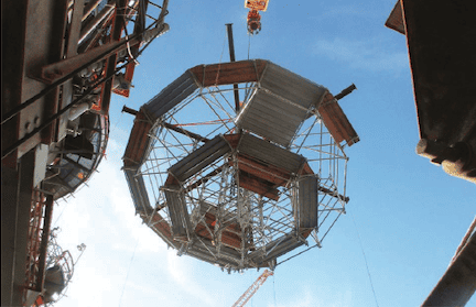 A large, circular metal structure is suspended mid-air by a crane, likely part of a project involving modular scaffold services. The octagonal framework appears to be integral to the construction. The sky is clear and blue, with surrounding construction elements partially visible at the edges of the image.