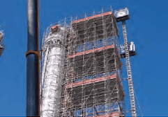 A building under construction is surrounded by professional scaffolding services. A blue crane is positioned nearby, and the clear blue sky is visible in the background. The intricate, extensive scaffold structure reaches several stories high, showcasing expert risk mitigation and abatement services.