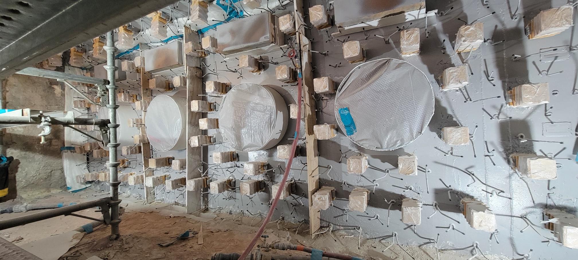 A concrete wall with multiple circular and square sections covered in protective wrapping, rebar ends protruding, and construction materials nearby.