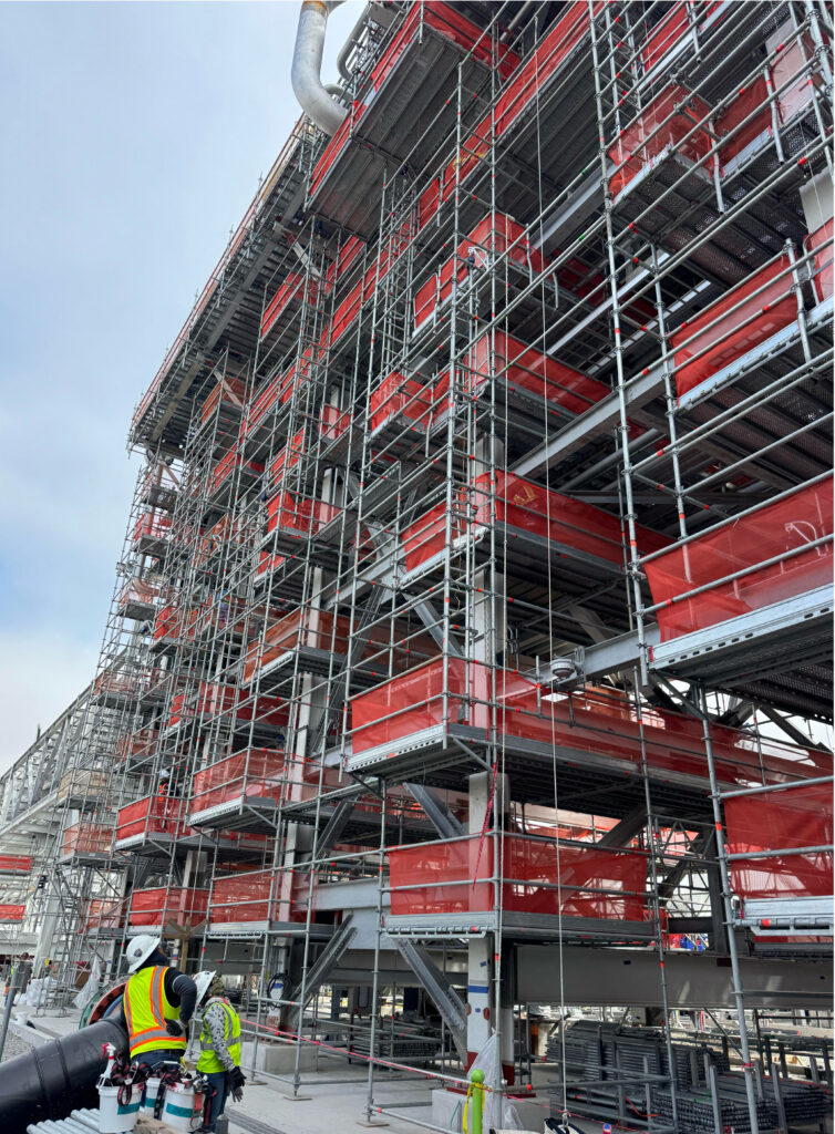 A large building under construction with extensive scaffolding and red safety netting; two workers in safety gear stand in the foreground.