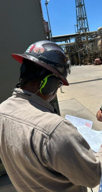 Person wearing a hard hat and ear protection reads Technical Support documents at an industrial site under a clear sky.