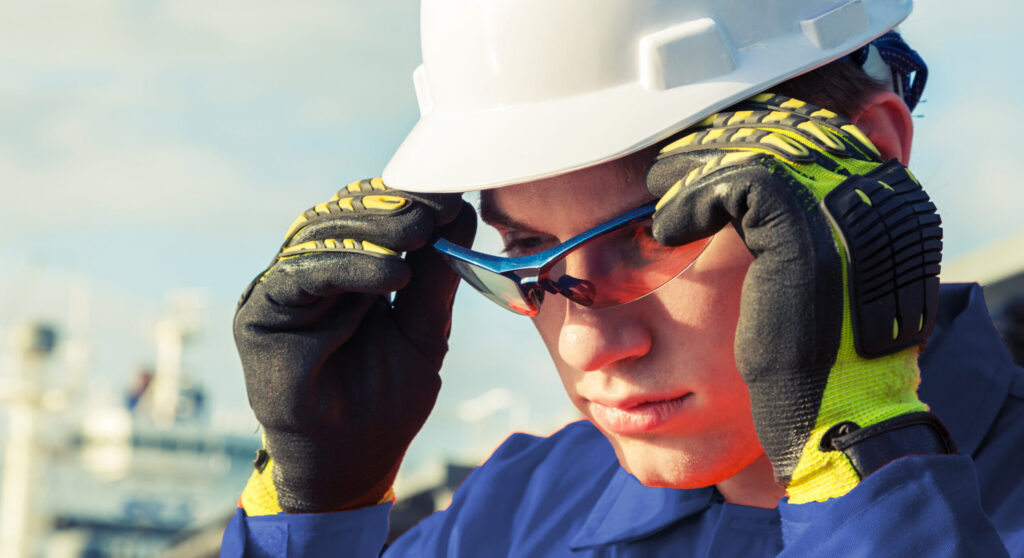 A worker in a blue uniform and white hard hat adjusts his glasses, ready to excel in safety at the industrial site.