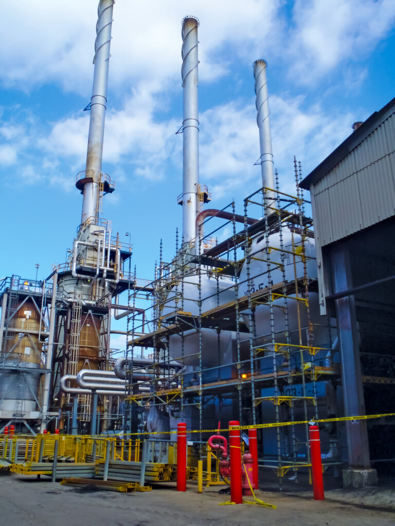 Industrial facility with tall metal smokestacks, scaffolding, and a team ensuring safety with caution tape and barriers under a partly cloudy sky.
