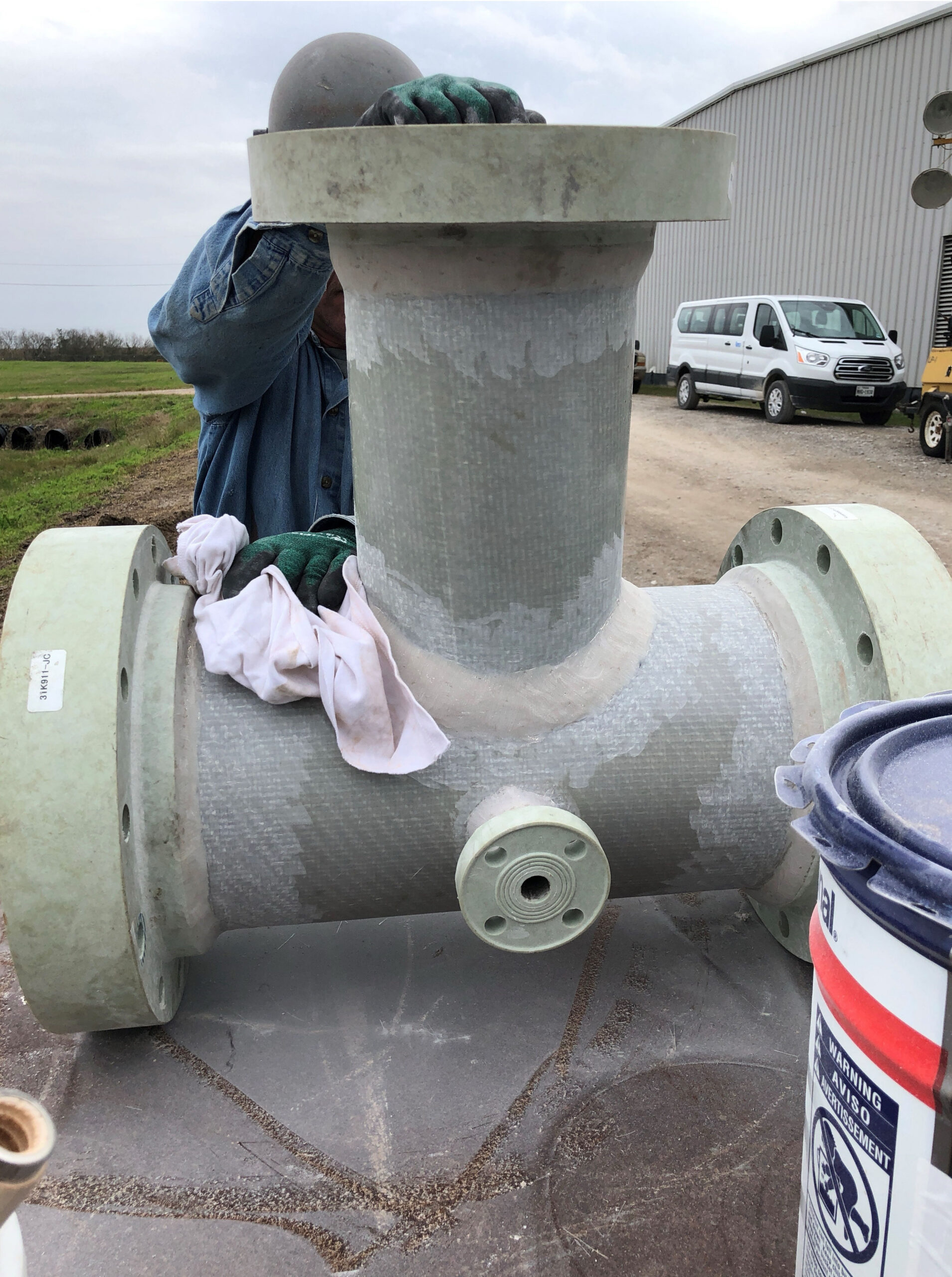 A person in gloves and a helmet showcases shop capabilities while working on a large T-shaped industrial pipe fitting outdoors.
