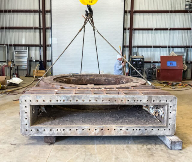 Large, rectangular metal structure with a circular opening showcases our shop capabilities, suspended by cables inside an industrial warehouse.