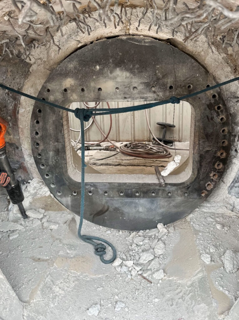 A circular metal hatch with bolts, partially open, surrounded by rubble and debris, showcasing shop capabilities with rope and tools visible.