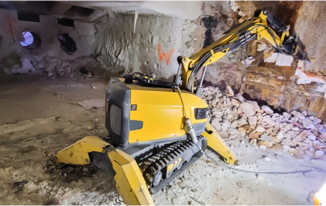 A yellow demolition robot with a hydraulic arm operates in a partially demolished, debris-filled room with exposed walls.